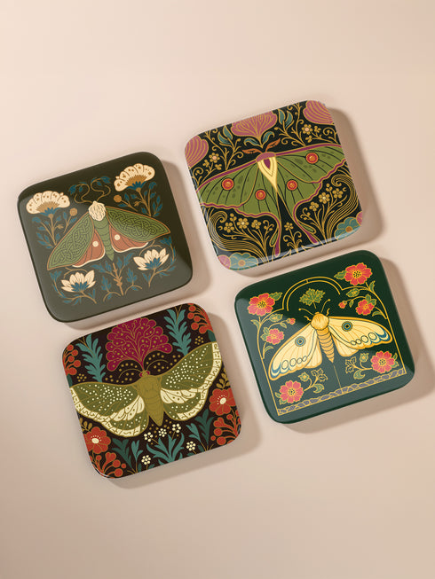 A set of four square hardboard coasters with smoothly rounded corners and a protective high-gloss finish. Each coaster features a unique, highly detailed Art Nouveau style illustration of a moth centered against a dark, botanical background. The intricate artwork uses a rich, moody color palette of deep forest greens, ochre, muted olive, crimson, and black, evoking a vintage, scholarly feel. These decorative drinkware accessories are designed for coffee table and bar cart use.